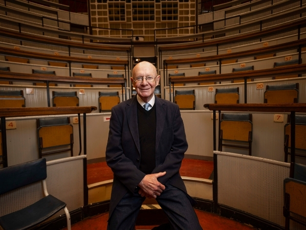Professor Ian Graham sitting in front of a lecture hall, smiling at the camera